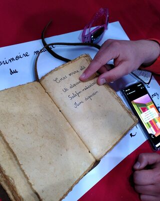 Une main tourne les pages du grimoire, à côté d’un téléphone qui affiche le jeu.